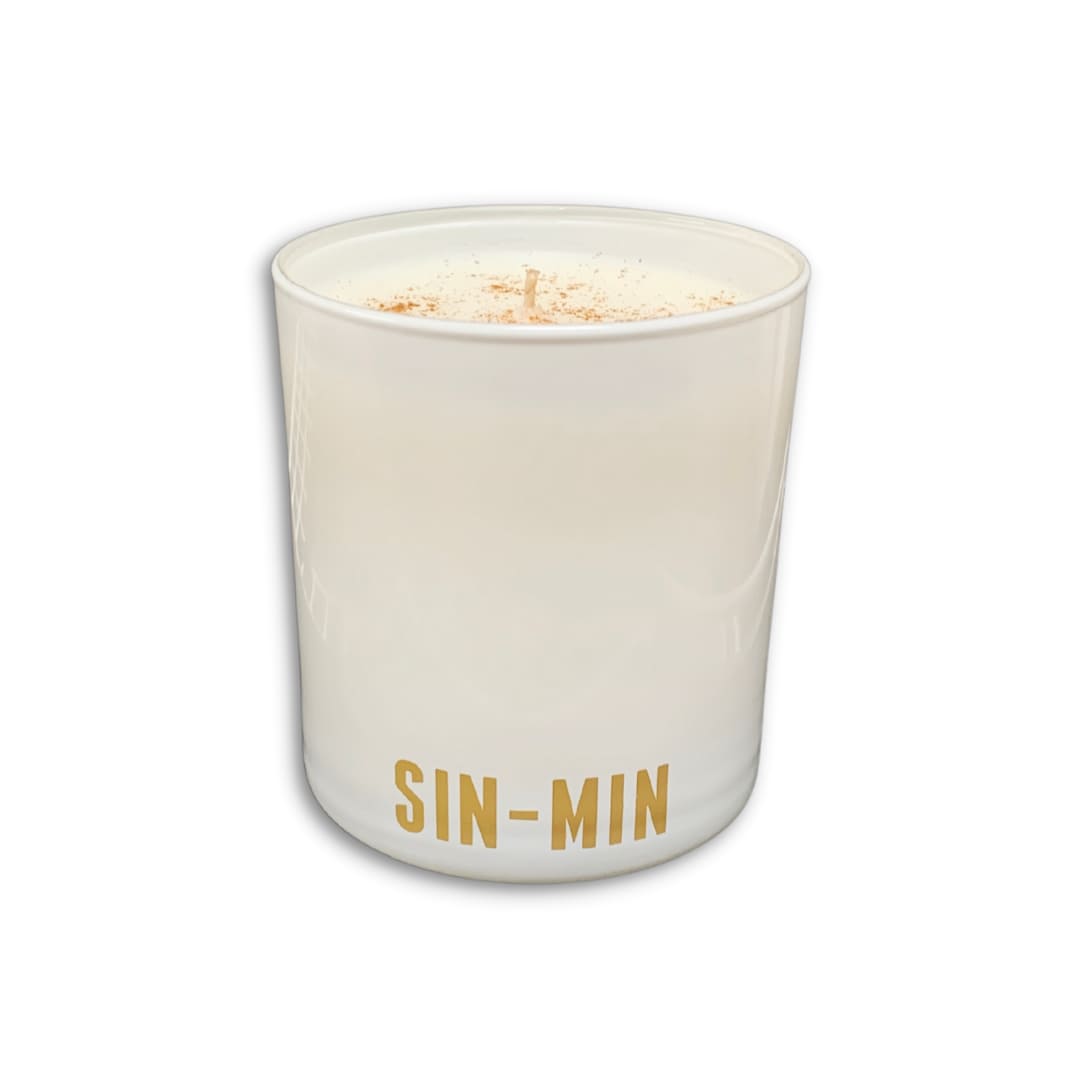 A white glass candle jar with the name 'SIN-MIN' printed in gold at the bottom, filled with white wax and topped with a light brown spice dusting, shown on a white background.