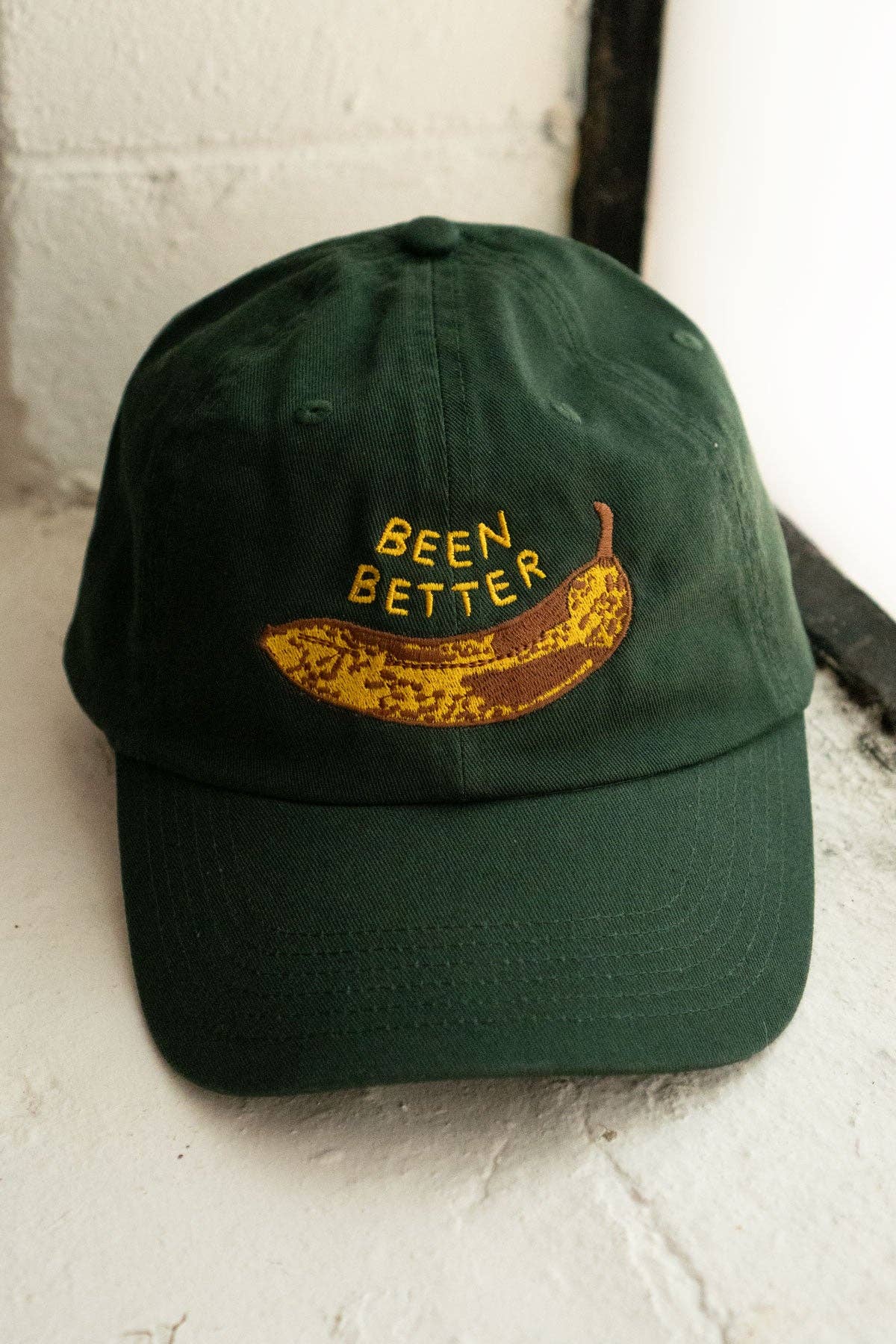 Forest green dad hat with an embroidered browned banana in the center with text above it that reads "Been Better"