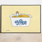 Wall print featuring an illustration of a girl sitting in a vintage CorningWare dish of Mac and Cheese with a pale yellow background. Shown in skinny black metal frame.