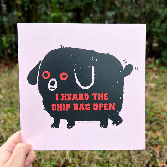 art print being held with image of a black dog with red eyes and red text that reads "I heard the chip bag open"