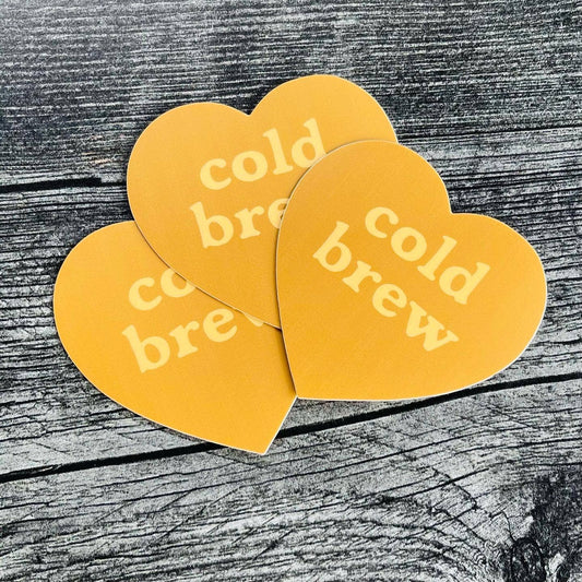 Three yellow heart-shaped stickers with 'cold brew' text on a wooden surface