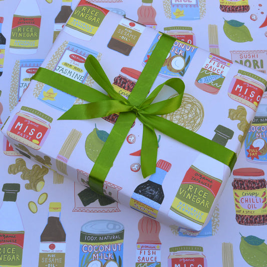 Gift wrap sheet with pale pink background featuring illustrated food items - Asian condiments and ingredients such as miso paste, rice vinegar fish sauce, sesame oil, chili oil, noodles,, nori seaweed sheets, lime and ginger. Shown on gift box with green ribbon.