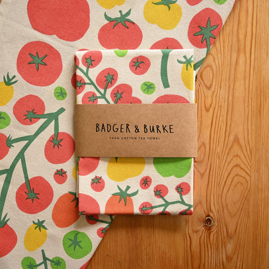 A folded cotton tea towel with a colorful tomato print is wrapped in a brown paper band labeled 'BADGER & BURKE'. The towel is displayed on a wooden surface next to a larger matching flat lay of the same towel.