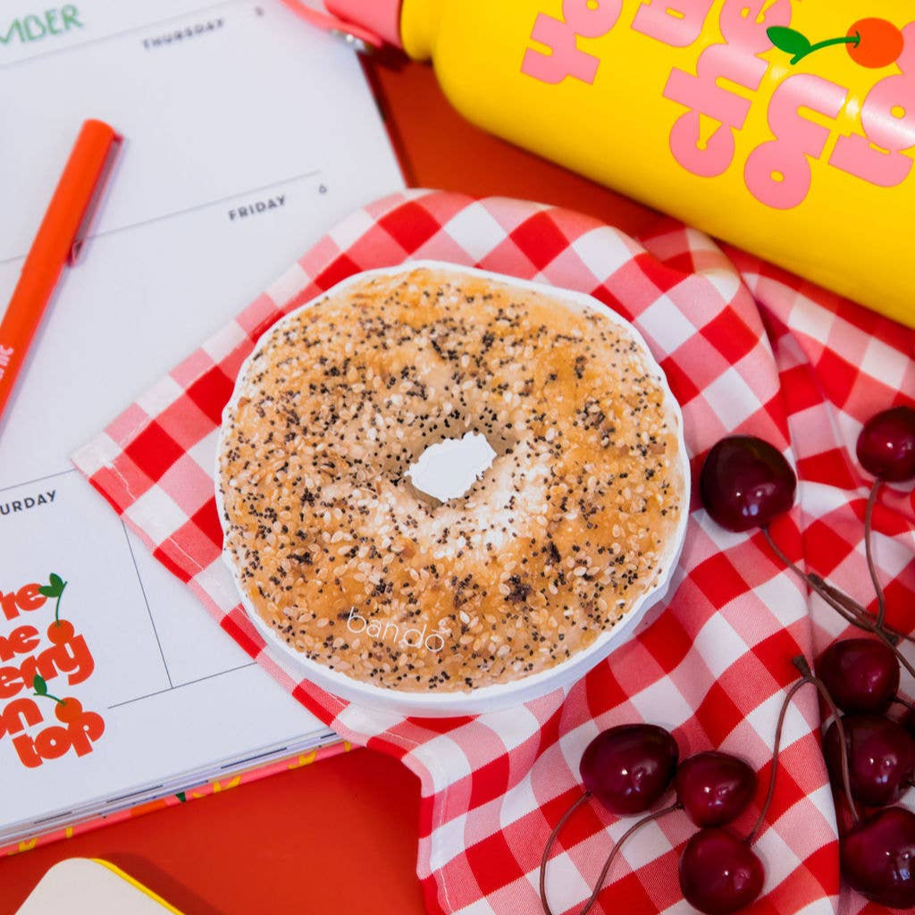 Bagel notepad with cream cheese on a checkered cloth with a yellow water bottle and cherries.