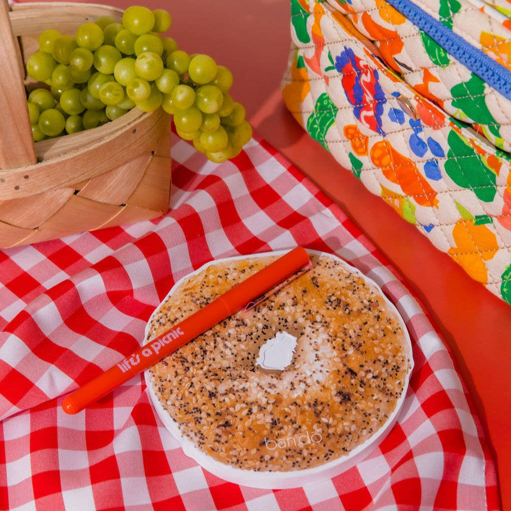 Bagel with cream cheese notepad on a red and white checkered tablecloth with a colorful cooler in the background