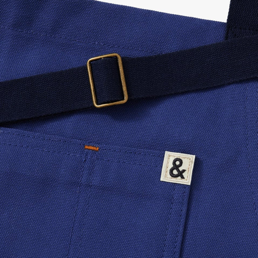 Blue apron with navy/black straps. This apron highlights 4 different styles of bagels. This picture shows a close up of the small ampersand logo on the pocket.