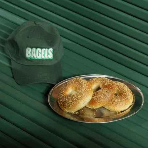 Dark green baseball cap with embroidered patch that reads "BAGELS" on the front. Pictured here with 3 sesame bagels on a silver platter 