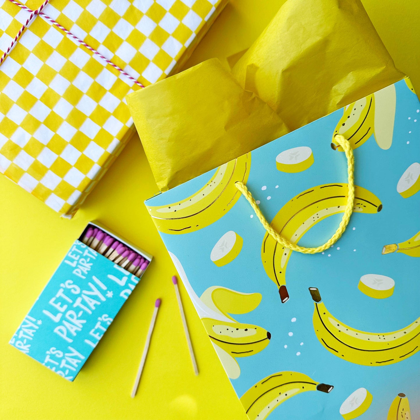 staged table top featuring a banana gift bag with yellow tissue. Checkered yellow tissue and matches on the table as well.