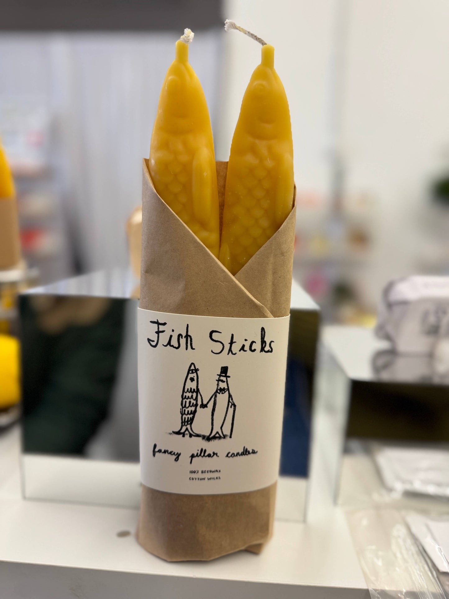 Set of two yellow beeswax taper candles shaped like fish. Shown in paper wrapping with "Fish Sticks" branding and logo.