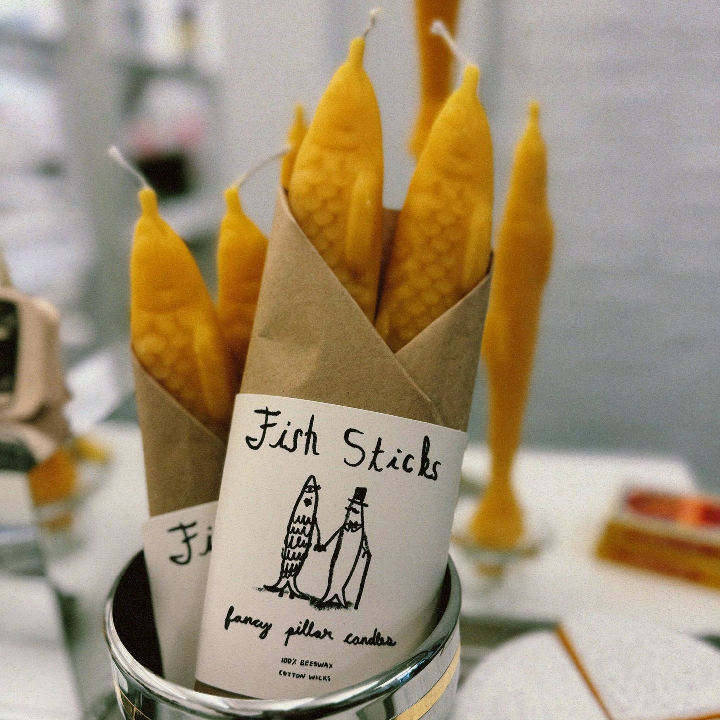 Set of two yellow beeswax taper candles shaped like fish. Shown in paper wrapping with "Fish Sticks" branding and logo.