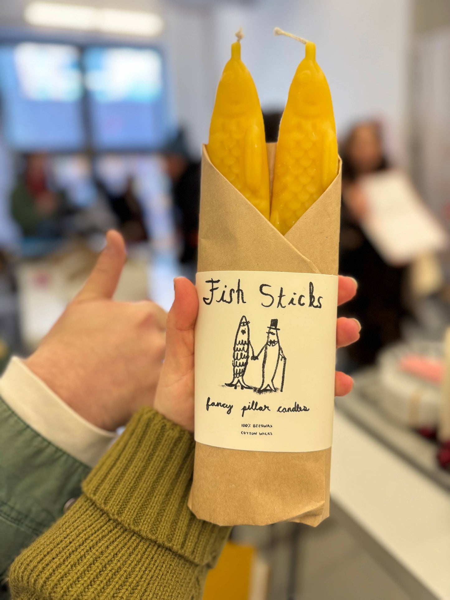 Set of two yellow beeswax taper candles shaped like fish. Shown in paper wrapping with "Fish Sticks" branding and logo. Package is held in a person's hand for scale.