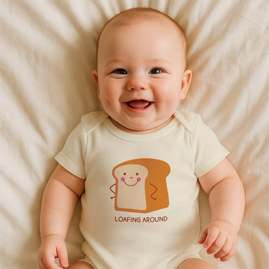 A smiling baby lying on cream-colored bedding, wearing a short-sleeve cream bodysuit with a cartoon toast graphic and the text 'LOAFING AROUND'.