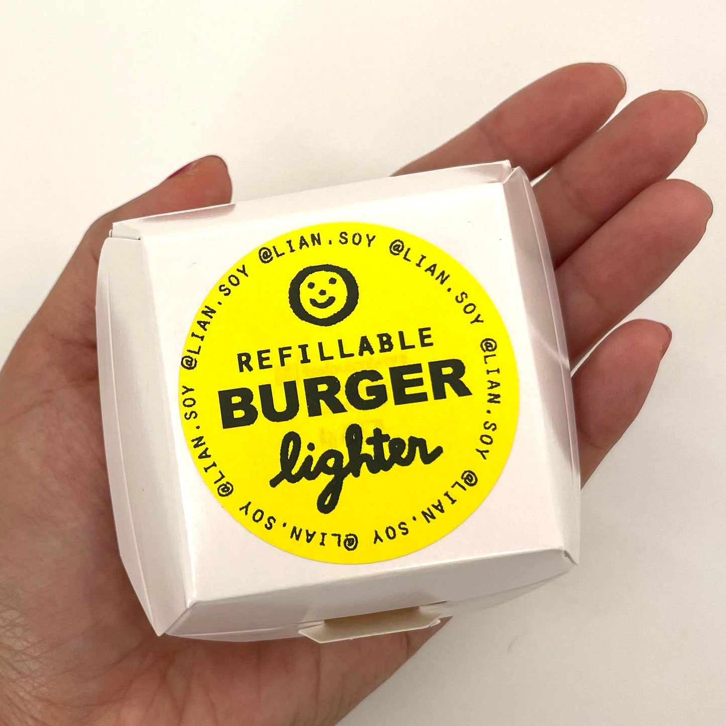 Packaging for burger lighter. Looks like a white takeaway box with yellow label "Refillable Burger Lighter" and instagram handle "@lian.soy"
