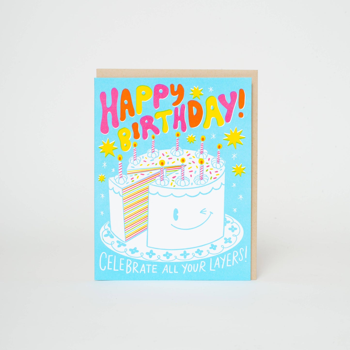 Greeting card with a winking birthday cake topped with candles - one slice is partially removed revealing many colorful layers inside. Blue background with yellow and white twinkles. Text reads "Happy Birthday! Celebrate all your layers!"
