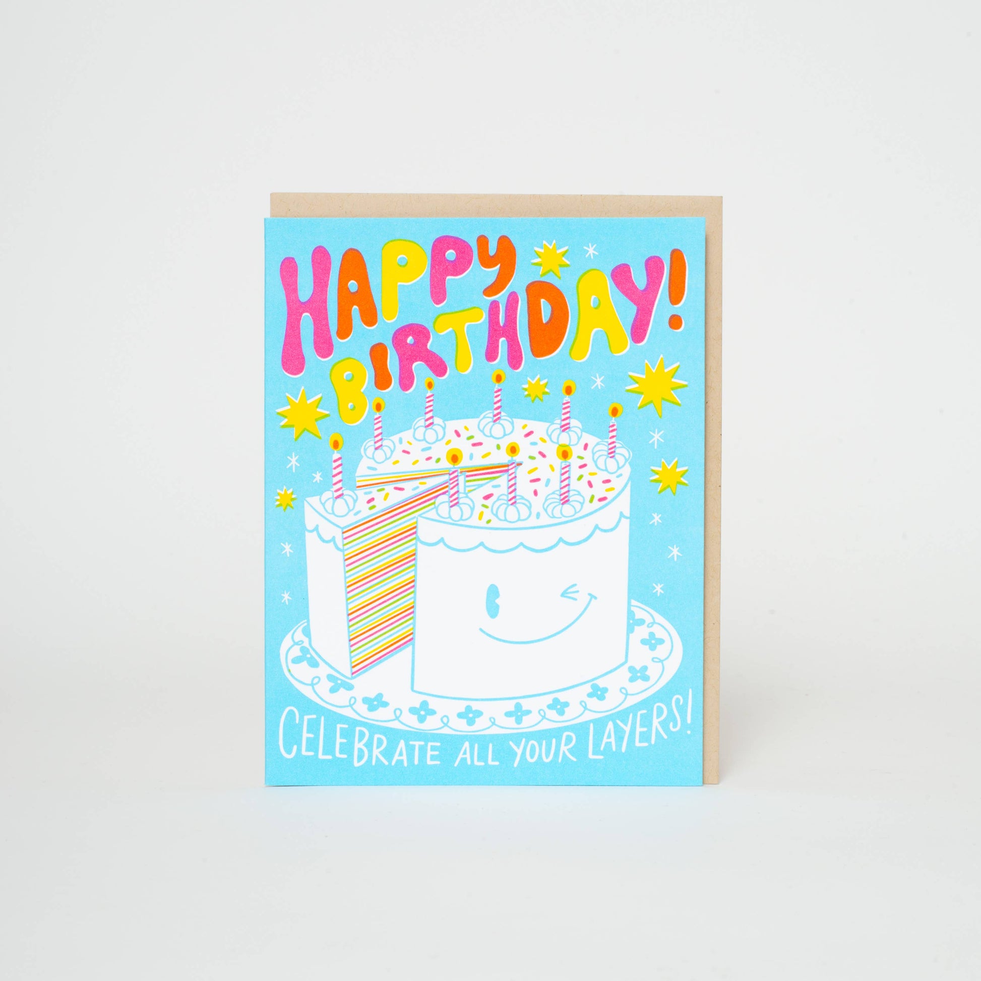 Greeting card with a winking birthday cake topped with candles - one slice is partially removed revealing many colorful layers inside. Blue background with yellow and white twinkles. Text reads "Happy Birthday! Celebrate all your layers!"