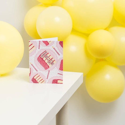 Greeting card with illustration of slices of pink birthday cake and candles on white background. Text "celebrating you is a piece of cake" is frosted onto the side of a piece of cake. Shown displayed on a white table with yellow balloons in the background.