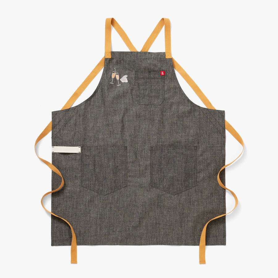 grey apron with embroidered champagne flutes filled with bubbles clinking. A single embroidered oyster in the shell next to it. Full sized layout of the apron to show the mustard colored straps and white towel loop.