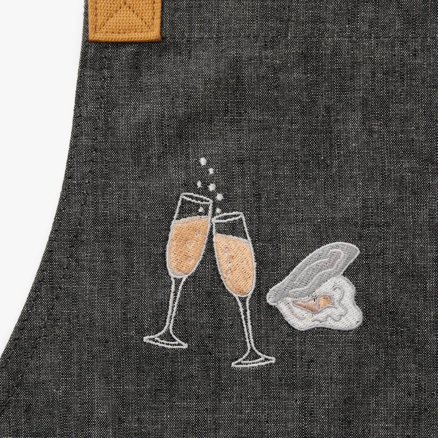 grey apron with embroidered champagne flutes filled with bubbles clinking. A single embroidered oyster in the shell next to it.