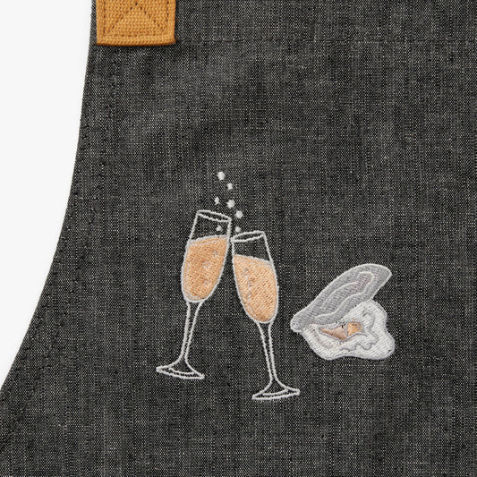 grey apron with embroidered champagne flutes filled with bubbles clinking. A single embroidered oyster in the shell next to it.