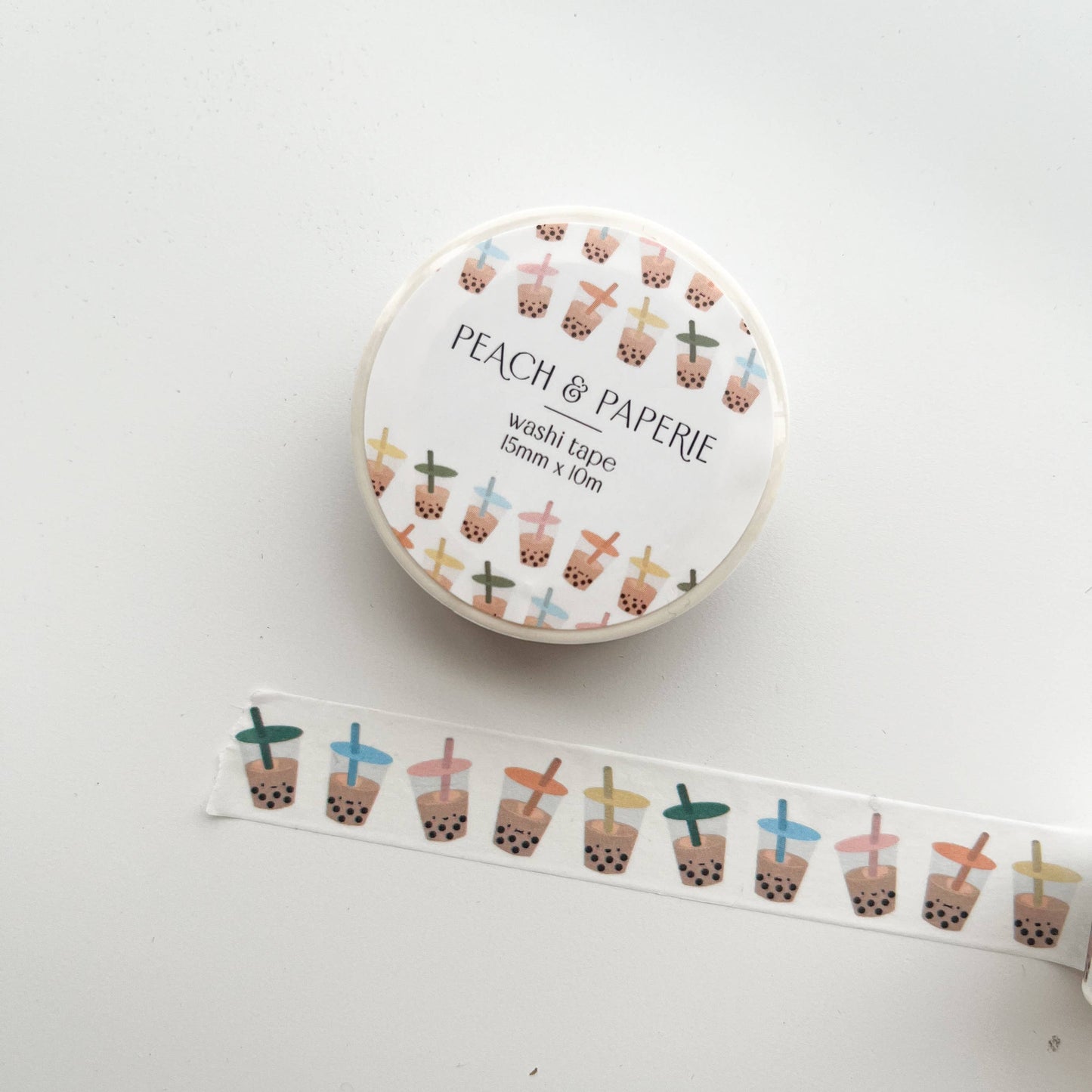 A roll of Peach & Paperie washi tape with a white label and a strip of the tape unrolled below, featuring a repeating pattern of boba tea cups in pastel colors on a white background.