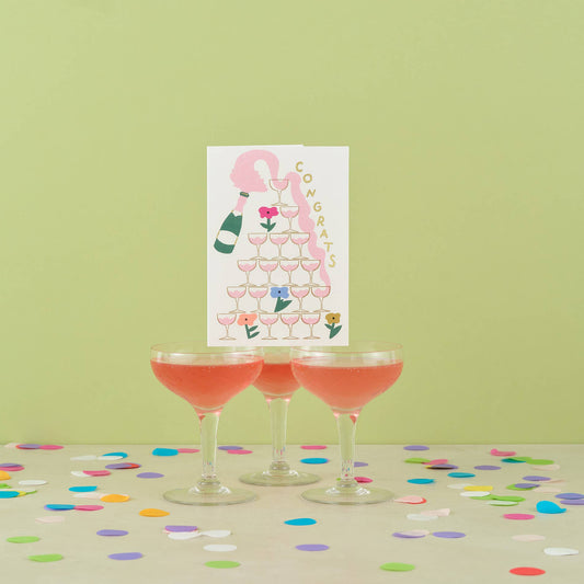 Three cocktail glasses with pink drinks on a table with confetti, next to a 'Congrats' card with champagne and flowers.