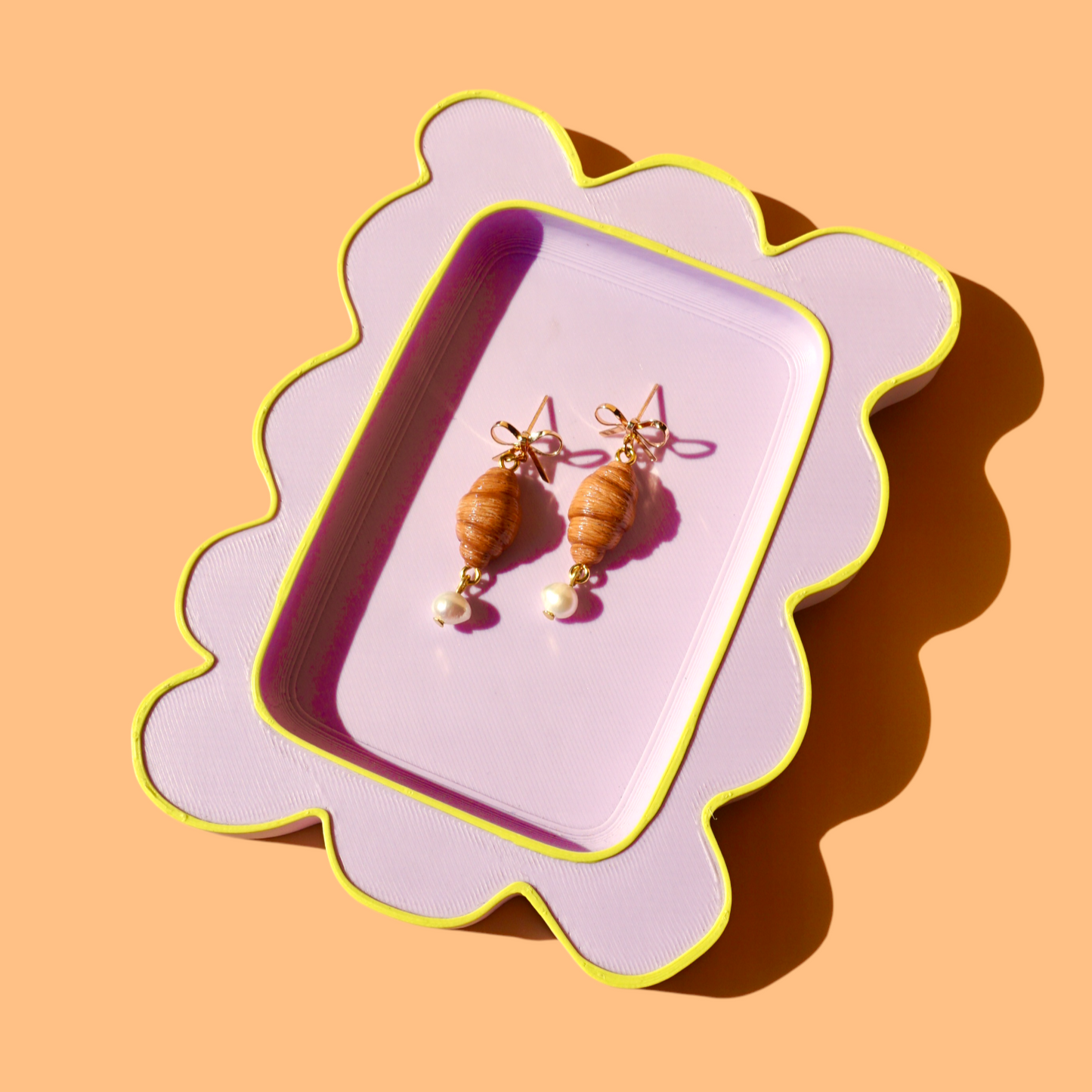 Pair of croissant earrings on a decorative pink and yellow tray against a peach background