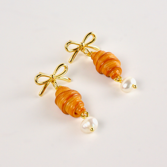 Pair of earrings with croissant-shaped beads and gold bows on a light background