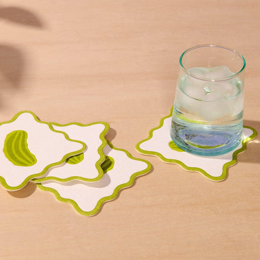 Three white coasters with wavy lime green edges are arranged on a light wood surface. One coaster is shown empty, and another is supporting a clear glass of water with ice.
