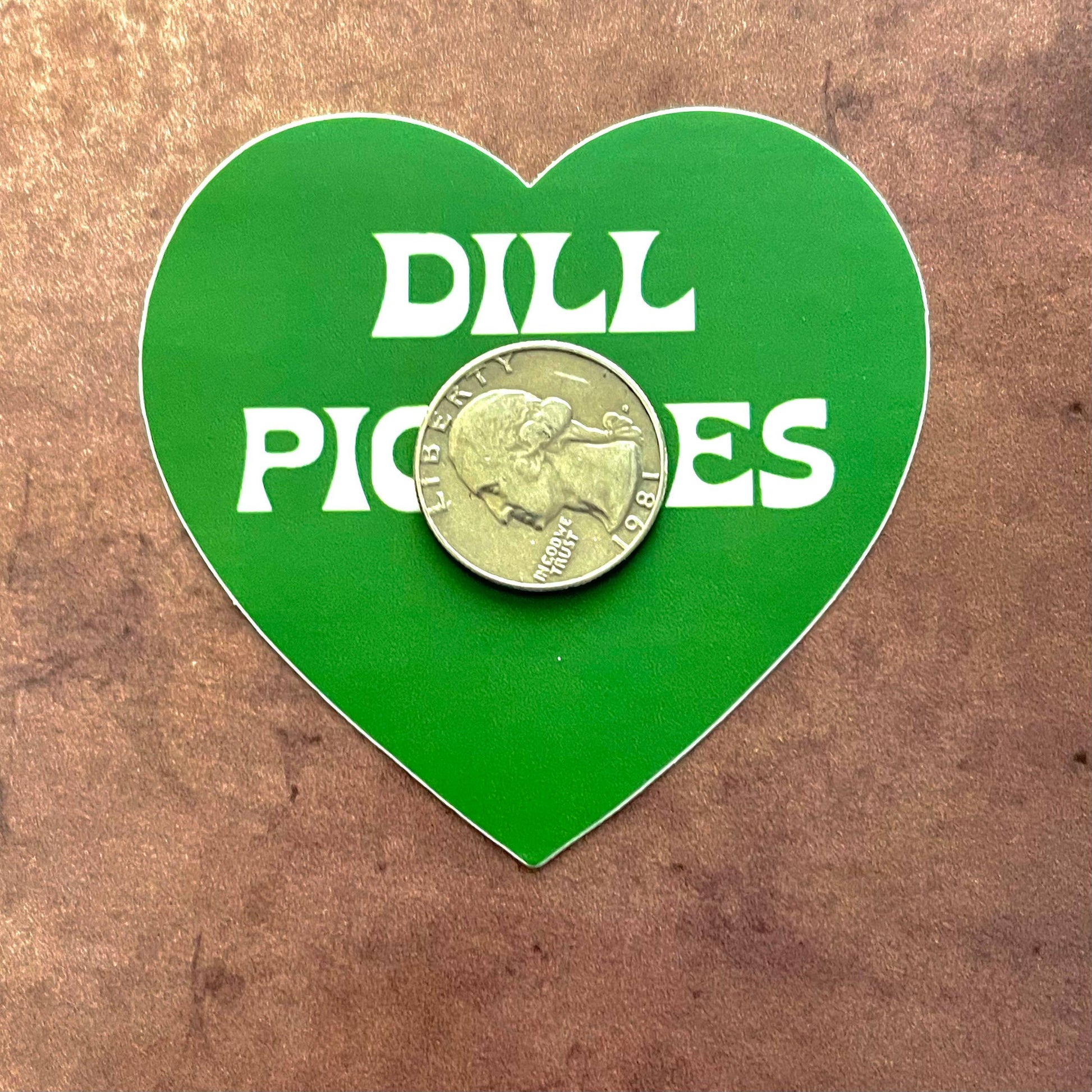 Green, heart shaped sticker with text in middle that reads "Dill Pickles"  with a quarter on top of it for scale