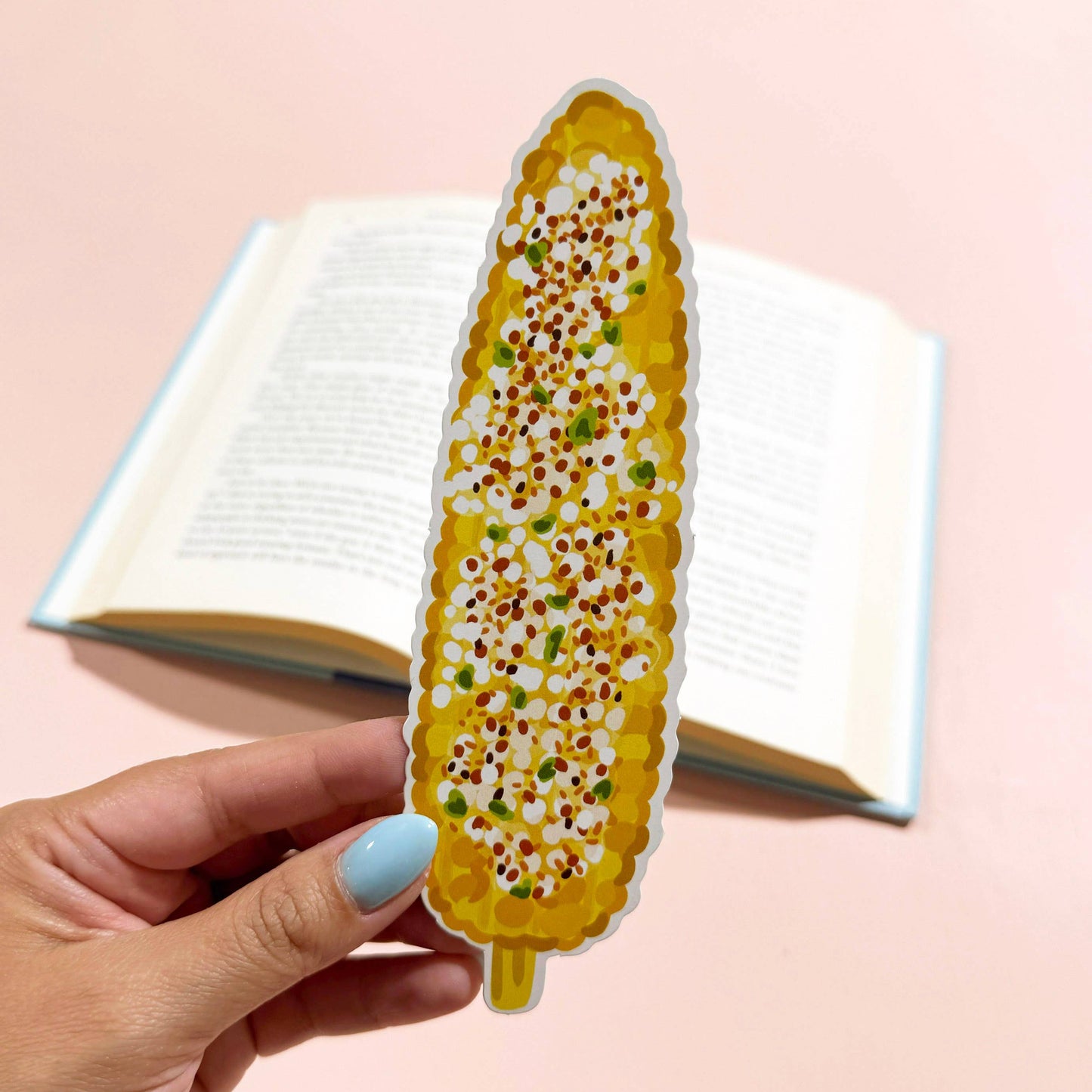 A hand with light blue nail polish holds a die-cut sticker shaped like a grilled corn cob with cotija cheese and tajin, positioned in front of an open book on a solid pastel pink background.