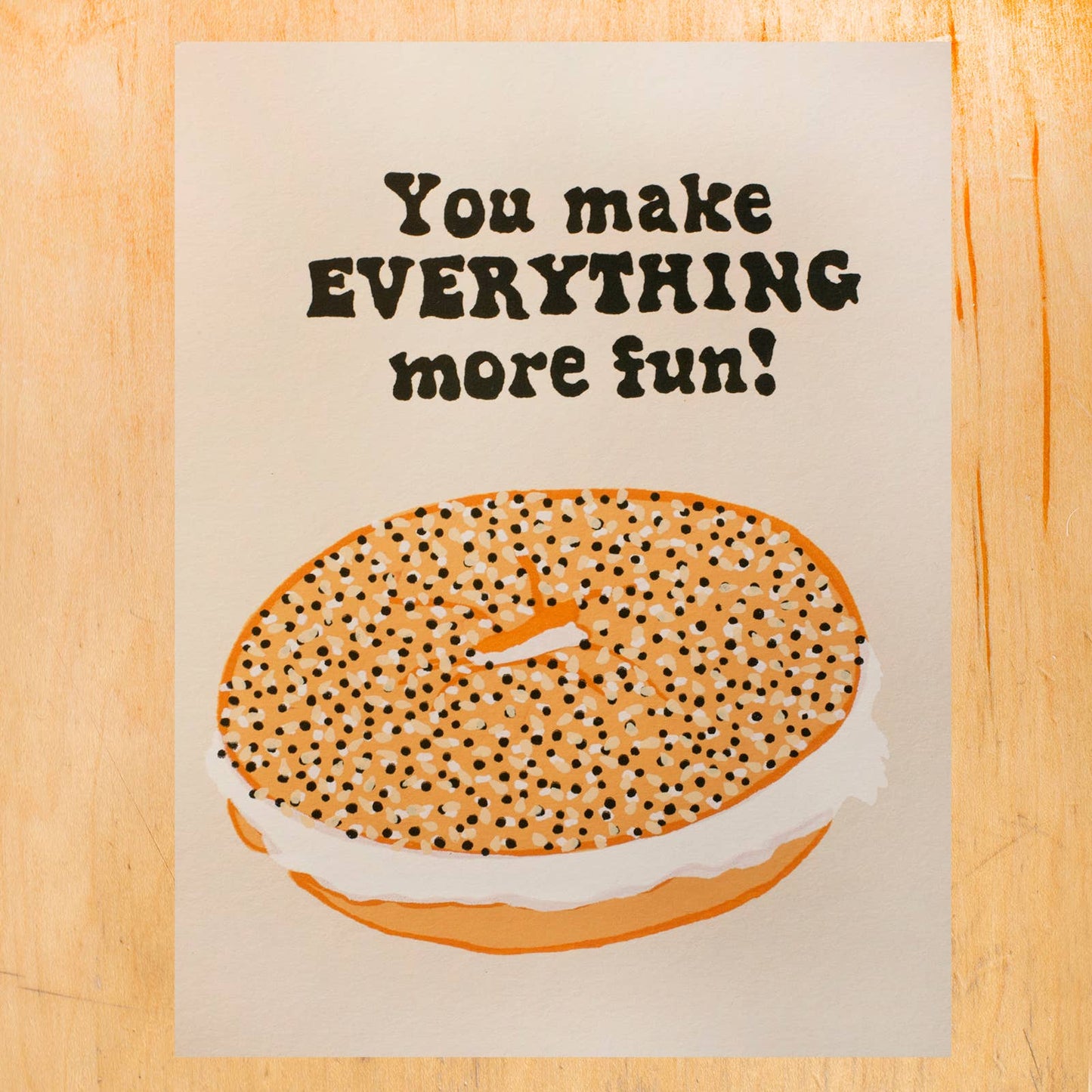 Card with a bagel illustration and text on a wooden surface. Text reads "You make everything more fun!"
