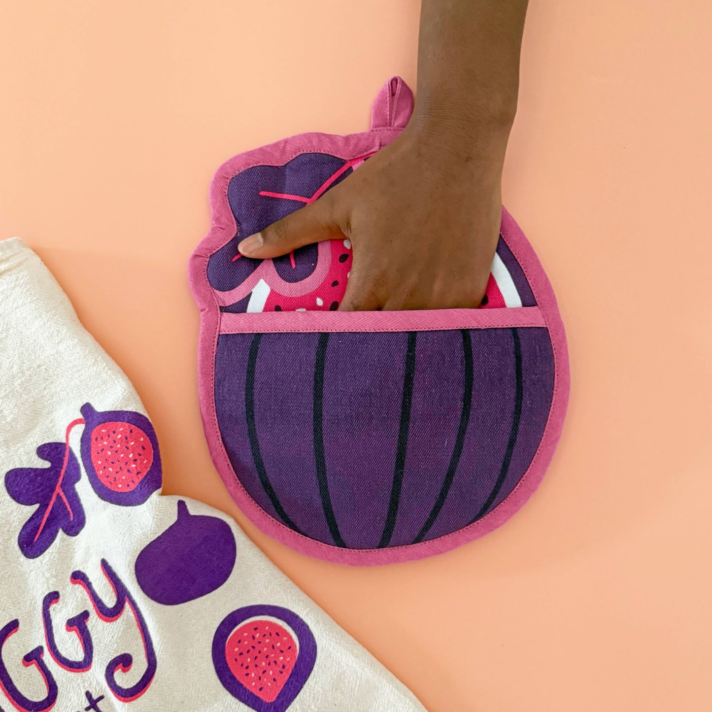 Purple and pink fig-shaped oven mitt held by a hand on a peach background with a white towel featuring fruit designs.