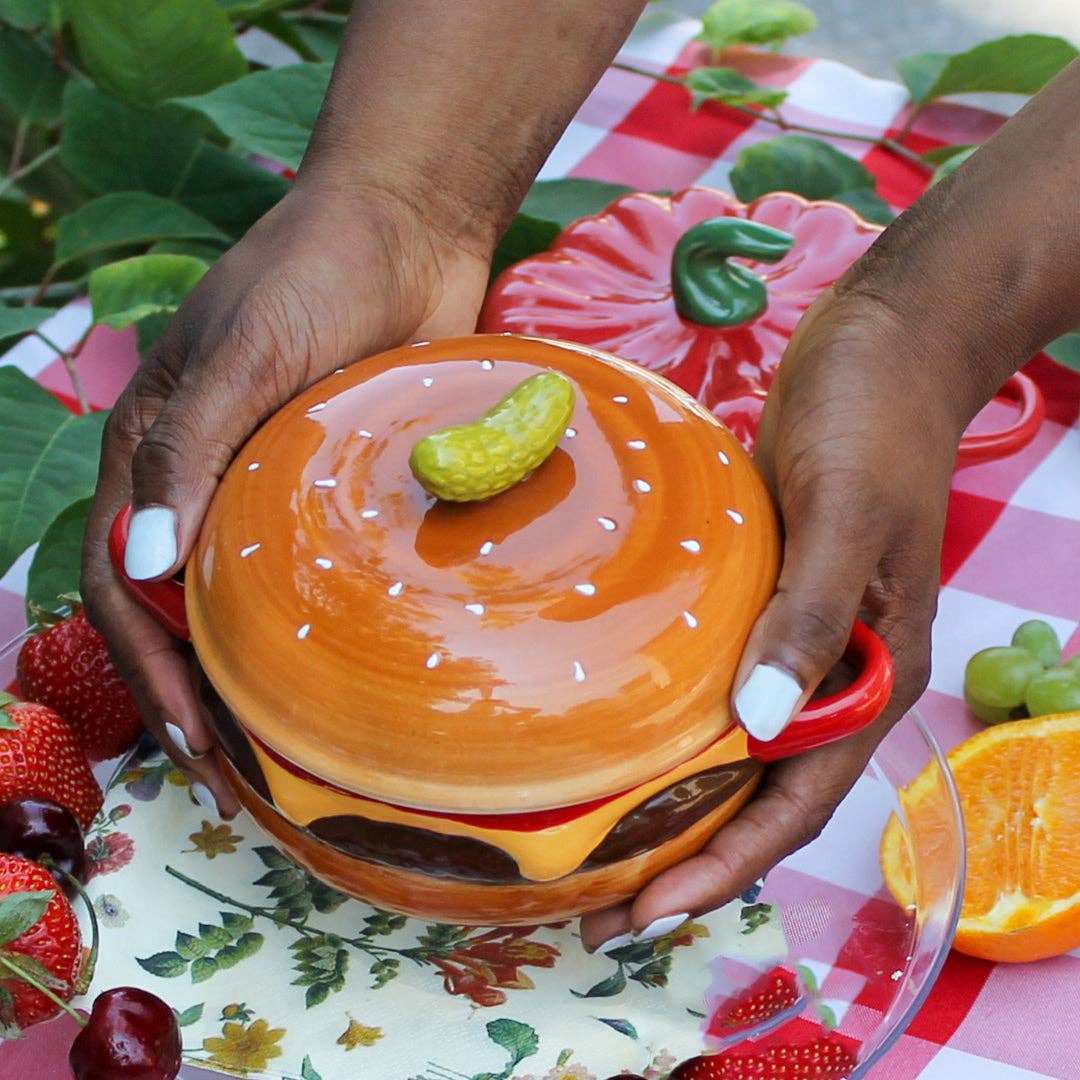 A ceramic cheeseburger-shaped bowl with red handles, held by two hands over a red-and-white checkered picnic tablecloth with strawberries, grapes, and an orange slice nearby.