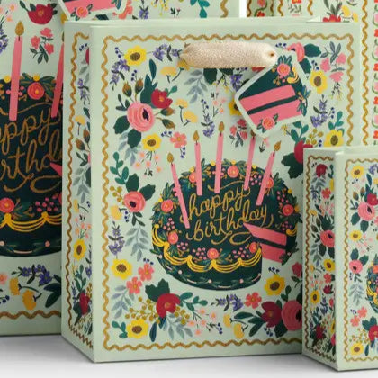 Gift bag shown in assorted sizes. Image on white background - intricate, vintage-inspired floral print surrounding birthday cake decorated with pink and purple flowers, dark frosting base, and pink candles. Squiggly golden yellow border. Very maximalist.