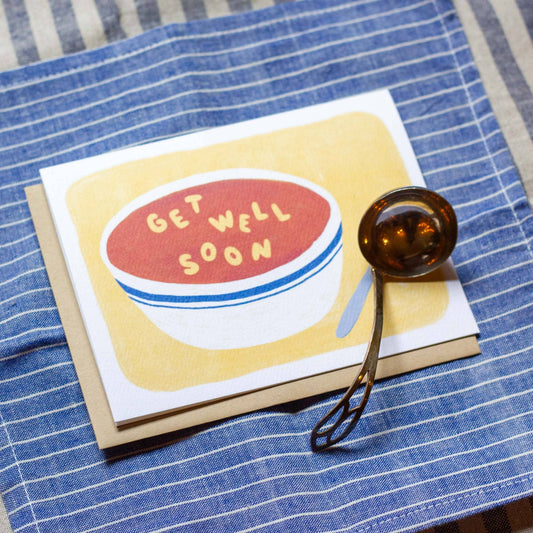 Greeting card with a bowl of soup with alphabet letters in it that spell out "get well soon" Bottom right of the card sits a ladle on top of the card
