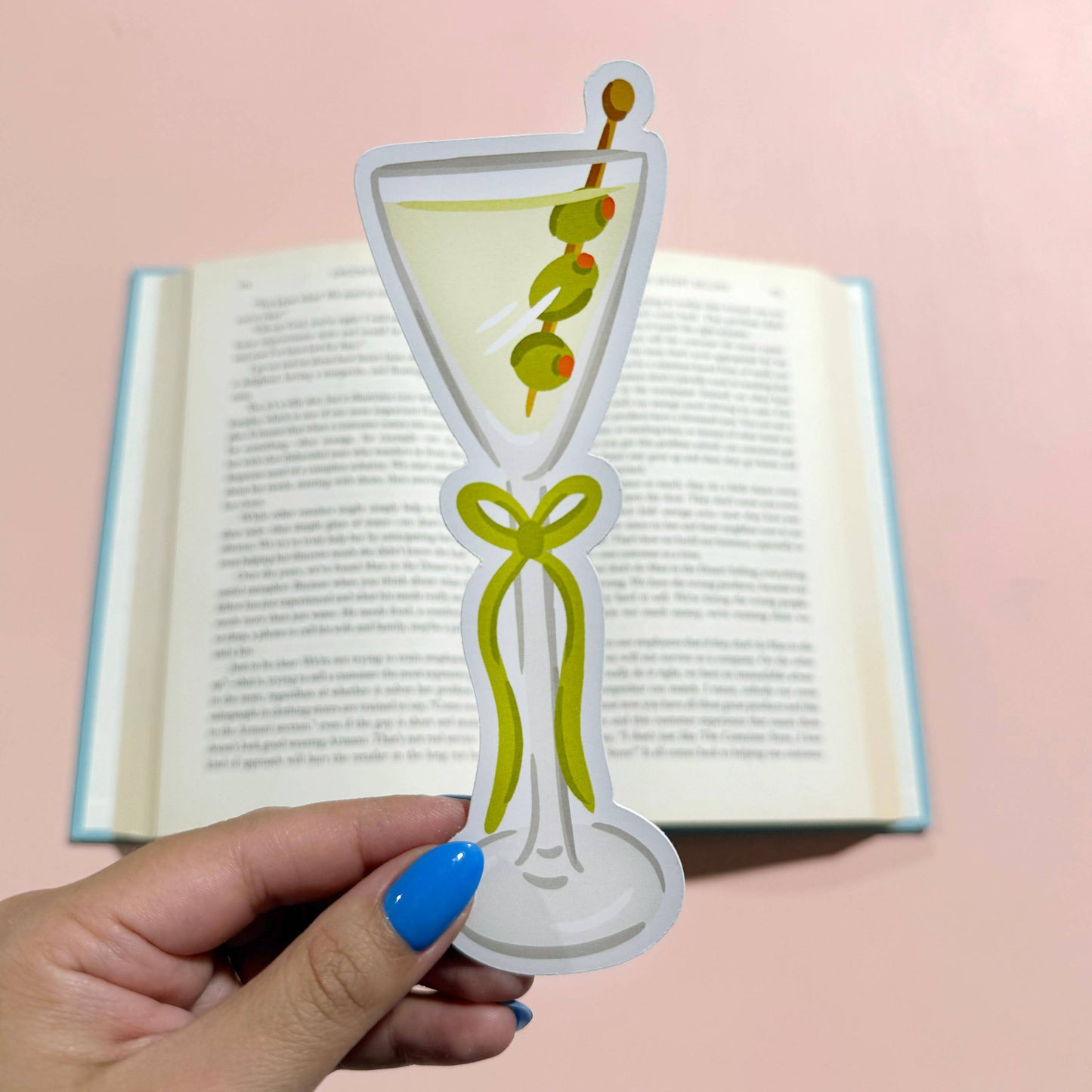 Hand holding a decorative bookmark shaped like a martini glass with olives on a pink background.