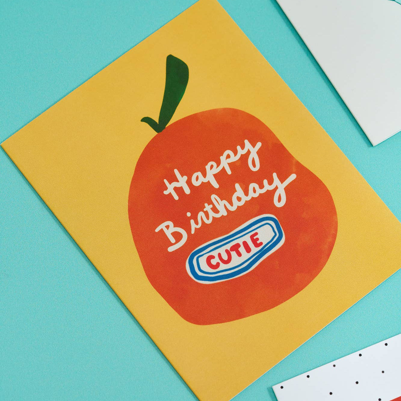 Birthday card with an orange design and 'Happy Birthday Cutie' text on a yellow background, placed on a turquoise surface.