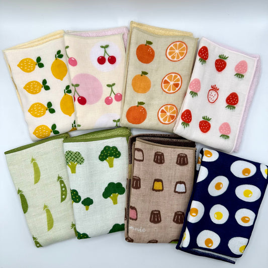 8 different hand towels folded and arranged in two rows. Designs include lemon, cherry, orange, strawberry, peas, brocolli, canele, and eggs. 