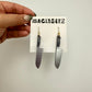 Hand holding a card with 2 earrings that look like knives. The handles are black and white checker. The card says Mackbecks.