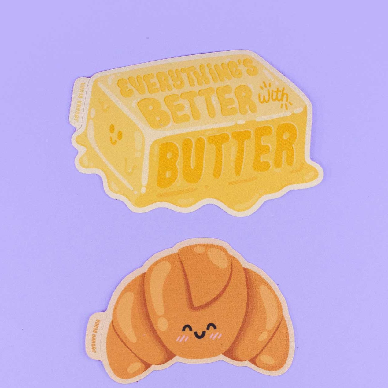 Two stickers on a purple backdrop. One of butter that says Everything's Better with Butter, and the second one that is of a croissant smiling.