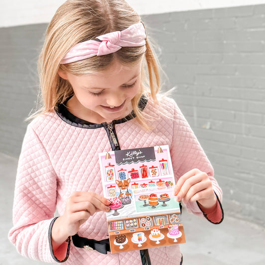 Interactive Card that includes stickers so one can decorate the front of the Sweet Shop with cakes and sweets. Text reads Kitty's Sweet Shop. Girl holding the card.
