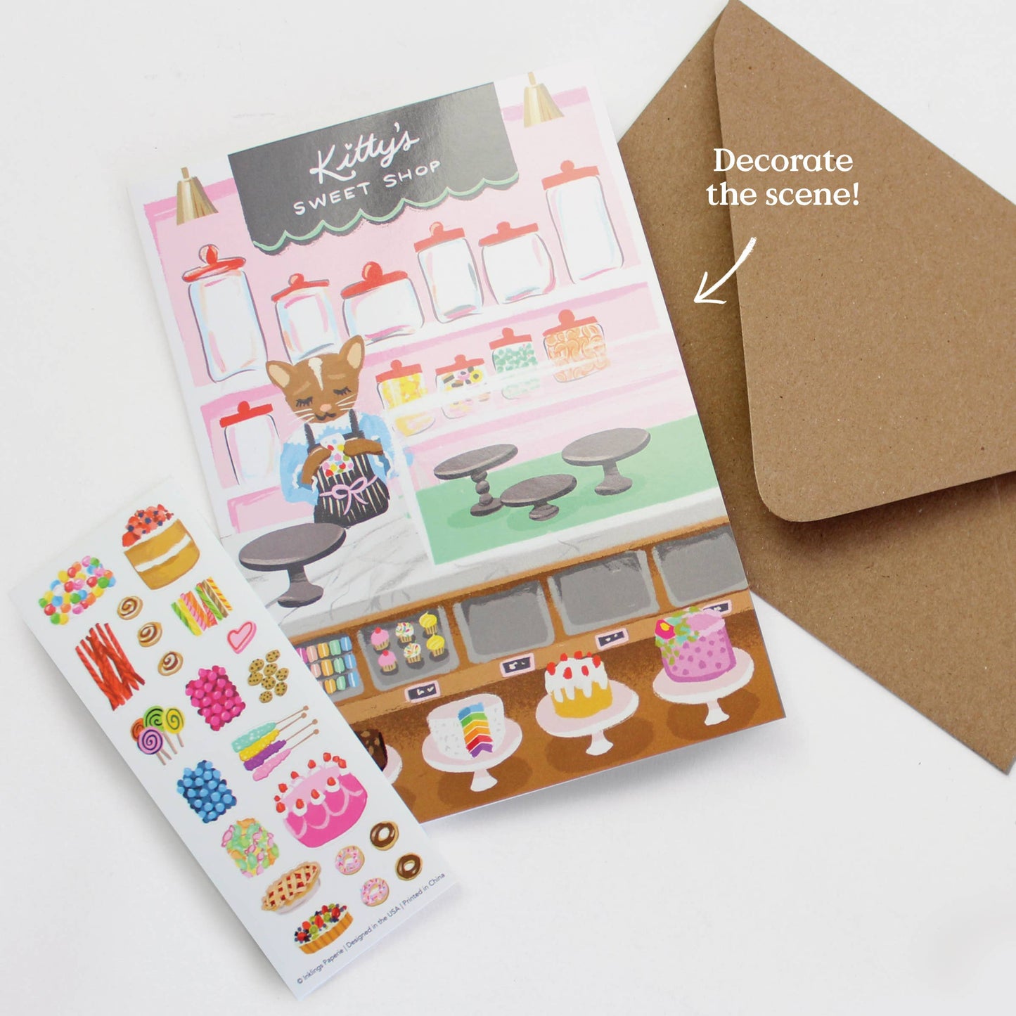 Interactive Card that includes stickers so one can decorate the front of the Sweet Shop with cakes and sweets. Text reads Kitty's Sweet Shop. Kraft envelope and directions that say "Decorate the scene"