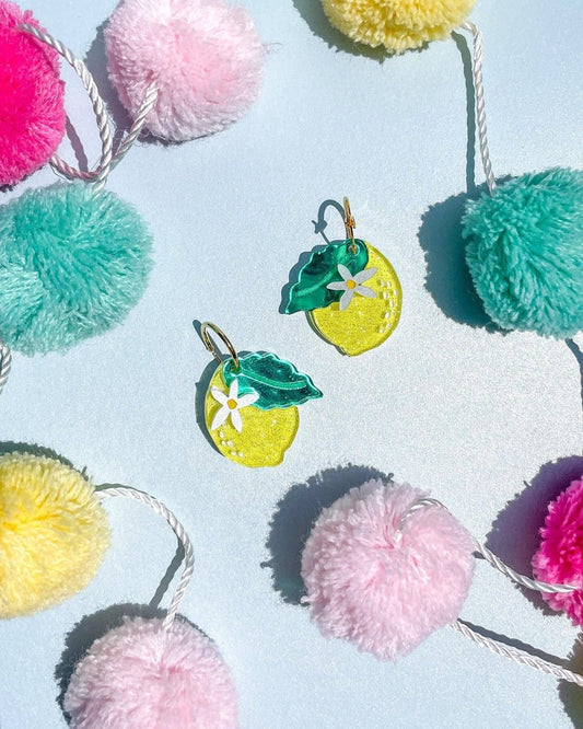 Acrylic earring shaped like a lemon with single green leaf and white daisy. Metal hardware. Shown with colorful pom poms for decor.