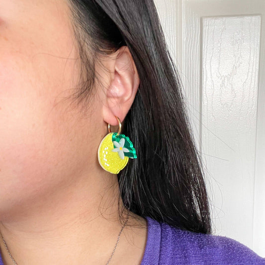 Acrylic earring shaped like a lemon with single green leaf and white daisy. Metal hardware. Shown worn on a person's ear.