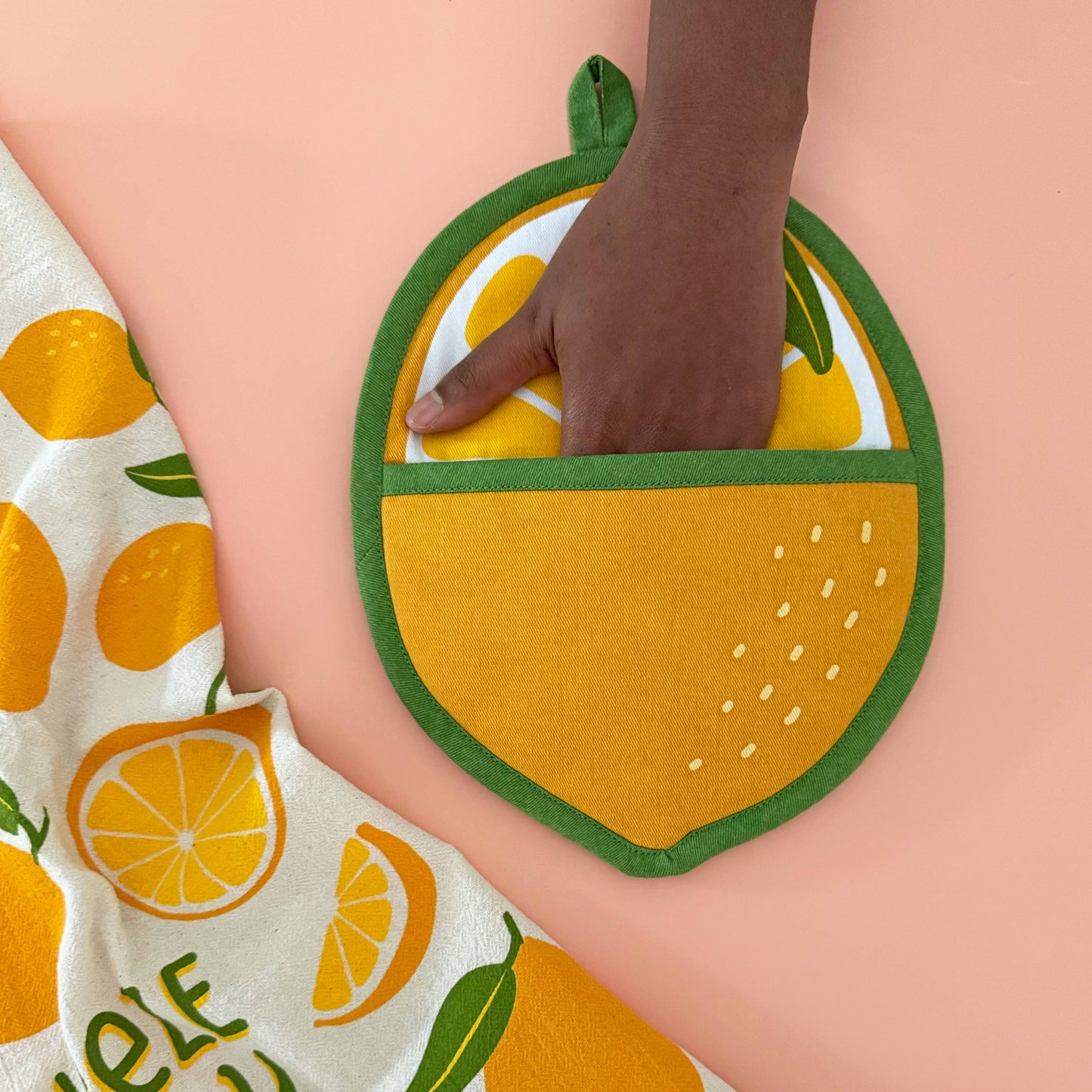 Hand holding a lemon-shaped potholder with a towel featuring lemon designs on a peach background