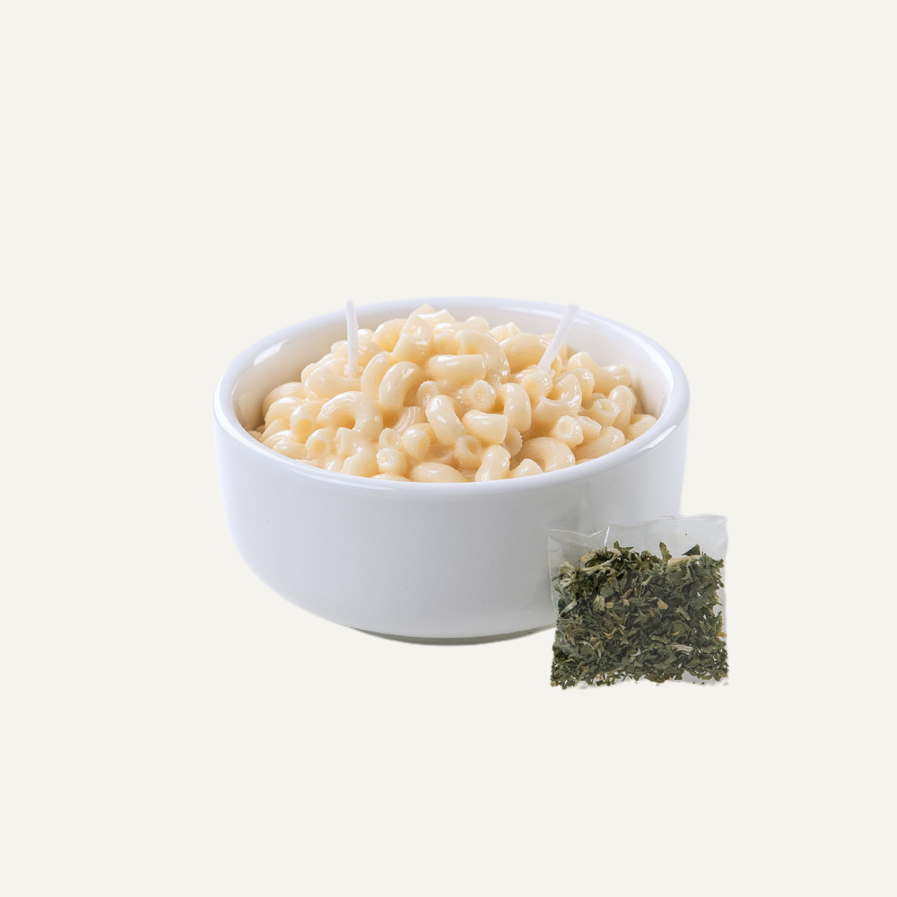 Candle that looks like a realistic bowl of white cheddar mac and cheese in a white porcelain bowl. Side of a baggie of herbs.