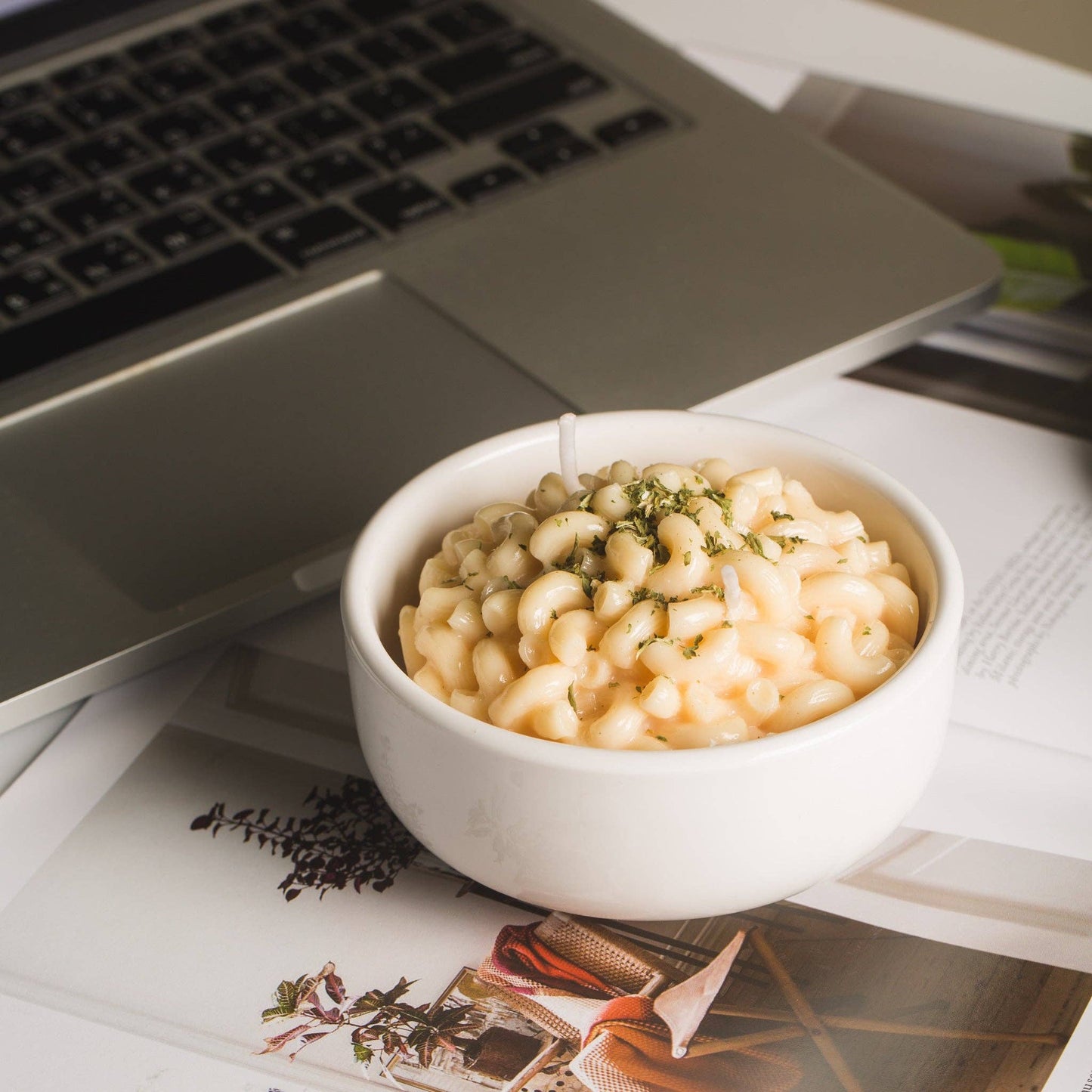 Candle that looks like a realistic bowl of white cheddar mac and cheese in a white porcelain bowl. Herbs sprinkled on top.Backdrop is a computer desk.