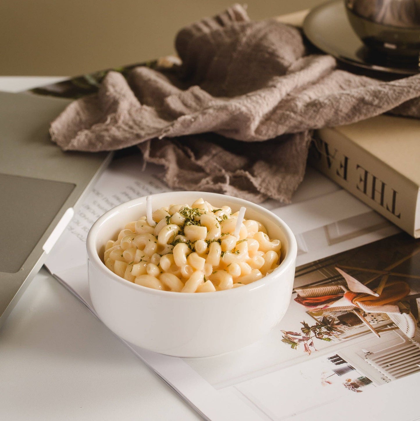 Candle that looks like a realistic bowl of white cheddar mac and cheese in a white porcelain bowl. Backdrop is a computer desk with magazines and a dish towel.