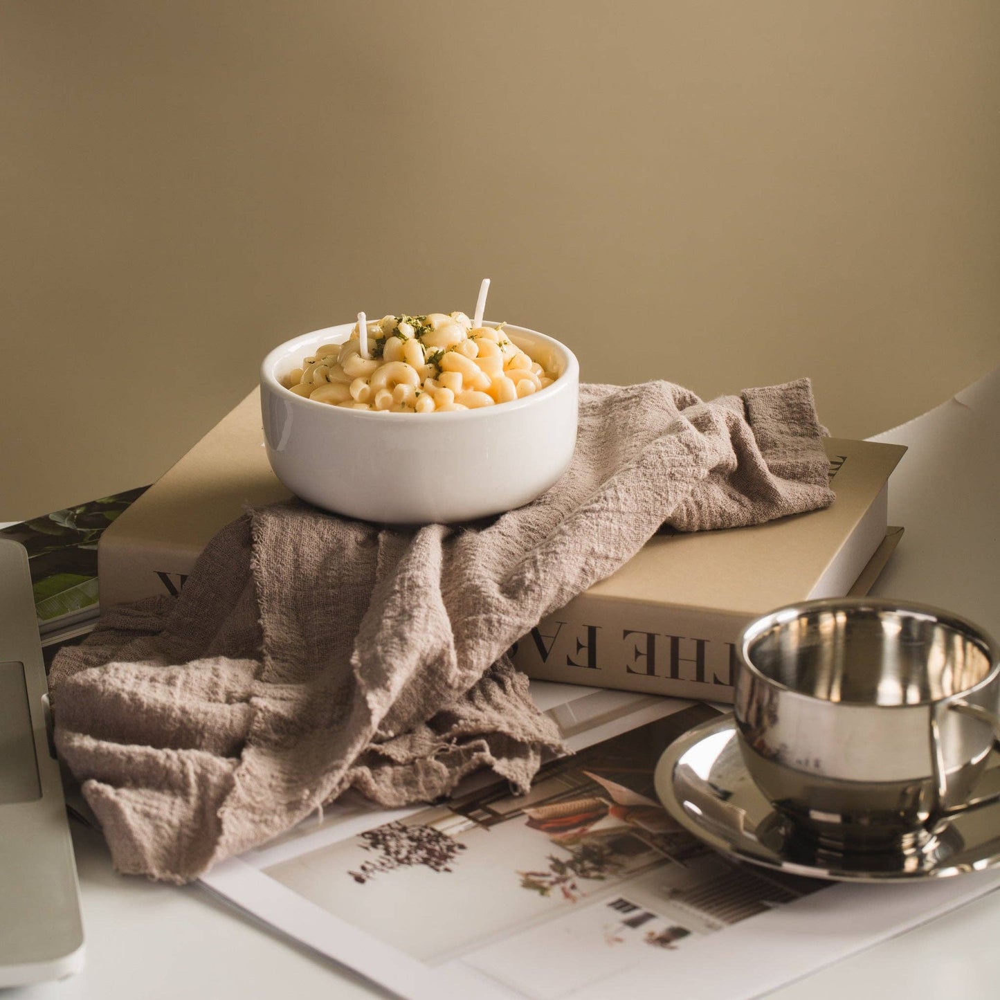 Candle that looks like a realistic bowl of white cheddar mac and cheese in a white porcelain bowl. Backdrop is a computer desk with magazines and a dish towel.