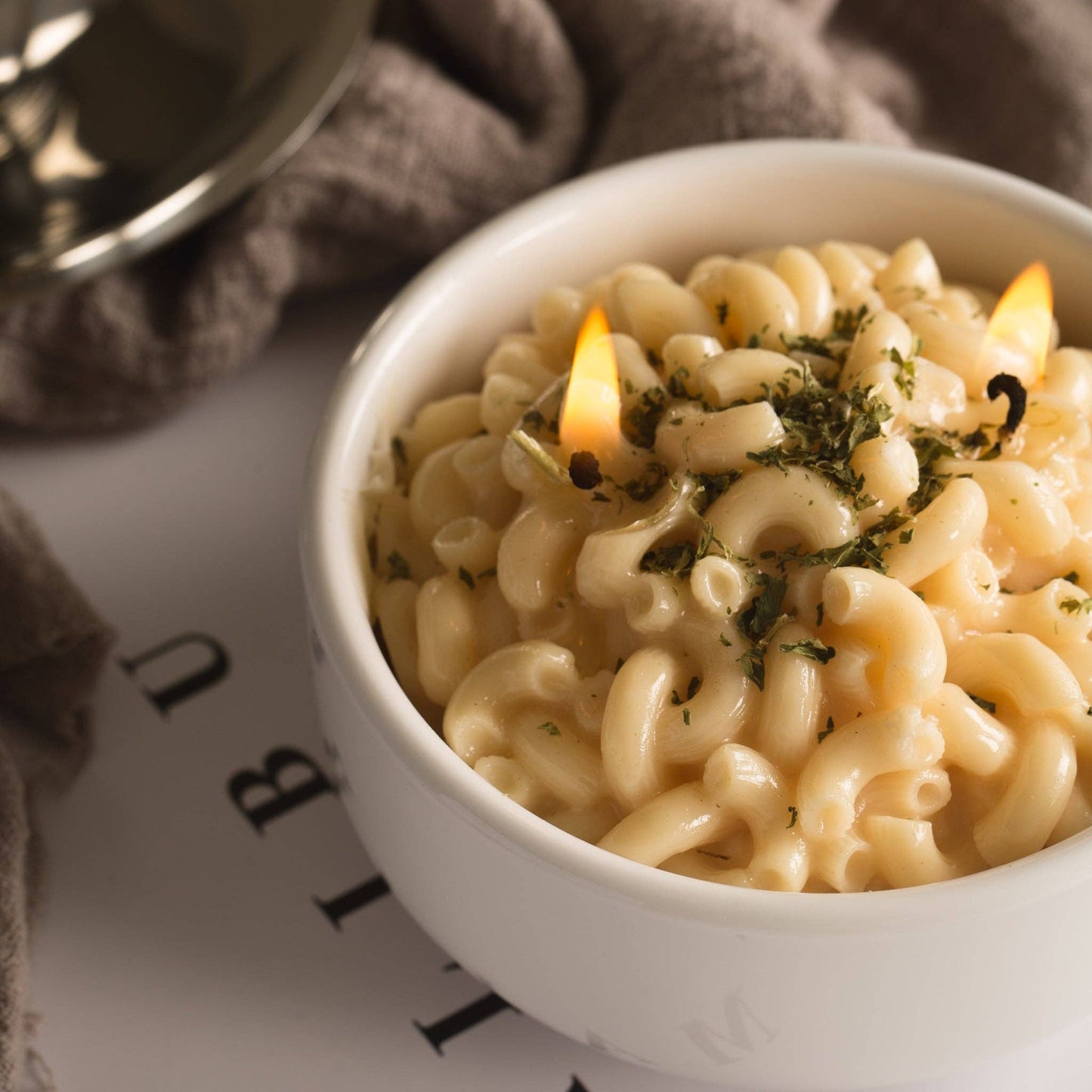 Candle that looks like a realistic bowl of white cheddar mac and cheese in a white porcelain bowl. Backdrop is a computer desk with magazines and a dish towel. Candle in this photo is lit.